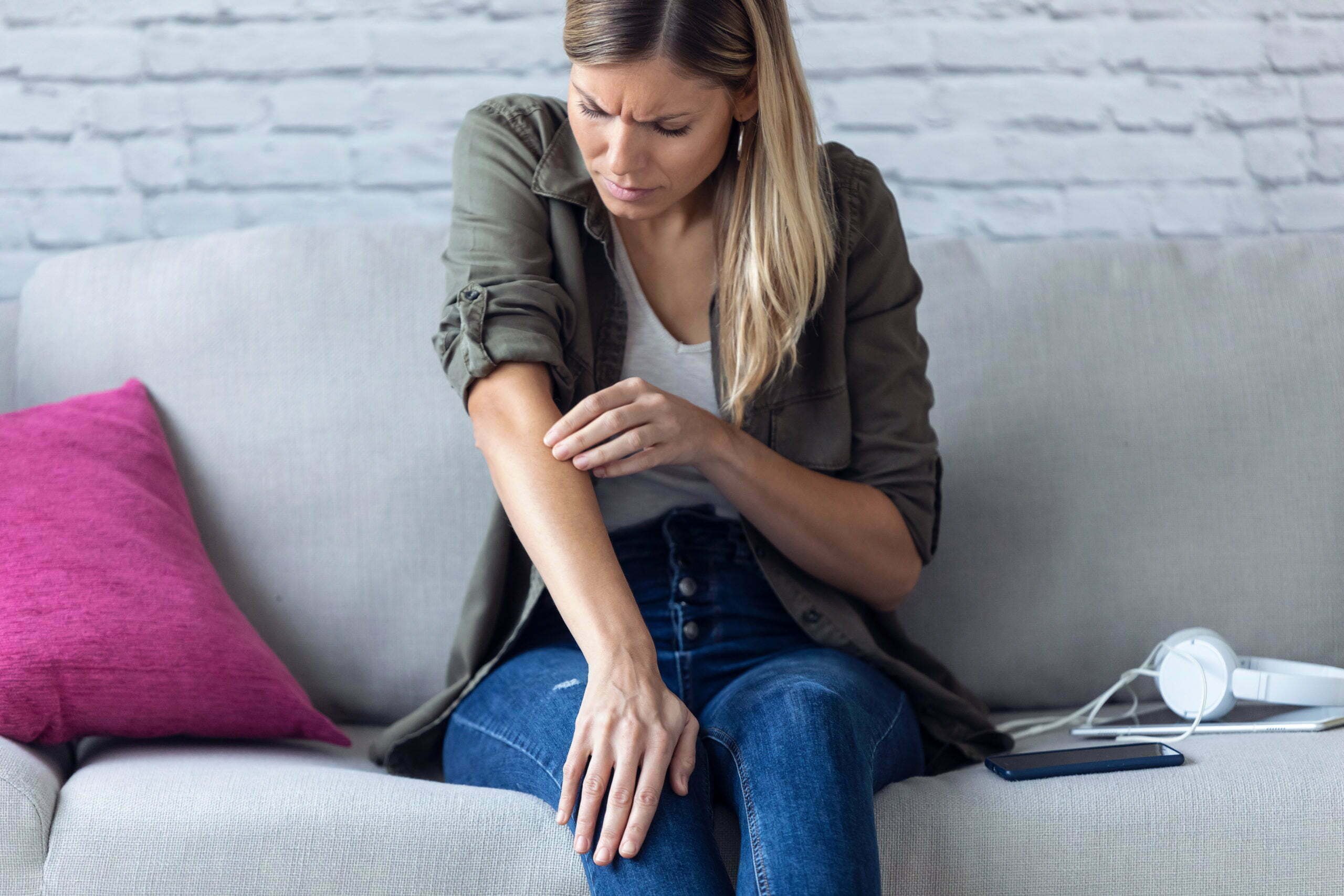 Uncomfortable young woman scratching her arm while sitting on the sofa at home.